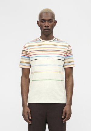 Mężczyzna z krótkimi blond włosami, ubrany w kolorową, poziomo paskowaną koszulkę z krótkim rękawem i ciemne spodnie, stojący na jednolitym tle.