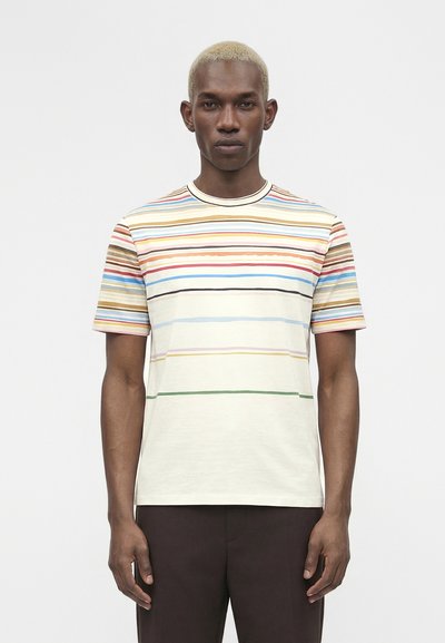 Man with short blonde hair wearing a multicolored horizontal striped short-sleeve shirt and dark trousers, standing against a plain background.