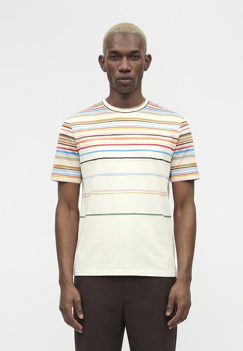 Άνδρας με κοντά ξανθά μαλλιά, που φοράει πολύχρωμο οριζόντιο ριγέ μπλουζάκι με κοντά μανίκια και σκούρο παντελόνι, στέκεται μπροστά σε απλό φόντο.