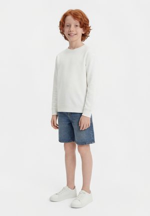 Noor punapõseline poiss, kes seisab ja naeratab, kandes valget pika varrukaga särki, teksapükse ja valgeid tossusid, taustal tavaline taust.