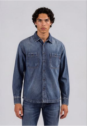 Giovane uomo con capelli ricci che indossa una camicia di denim blu sbiadita con due tasche sul petto e jeans blu, in piedi contro uno sfondo semplice.
