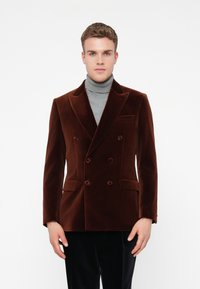 Vyras, vilkintis rudą aksominį dvigubą švarką virš pilkos golfinės, stovi priešais paprastą baltą foną.