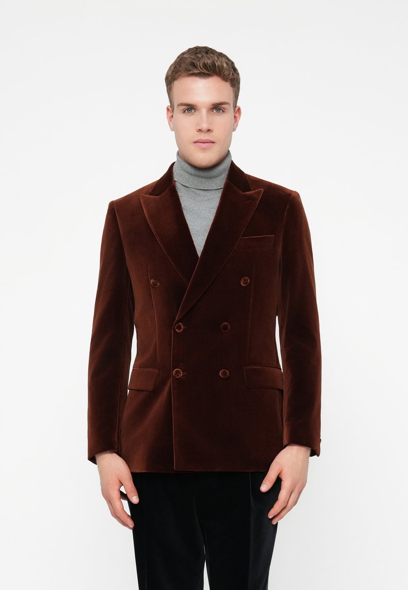 Vyras, vilkintis rudą aksominį dvigubą švarką virš pilkos golfinės, stovi priešais paprastą baltą foną.