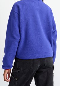 Giacca in pile viola con superficie texture, colletto alto e design corto, abbinata a pantaloni cargo scuri con tasche laterali.