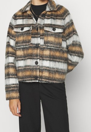 Persona que lleva una chaqueta de cuadros borrosa en tonos blancos, marrones y grises con botones negros y pantalones negros, de pie frente a un fondo liso.