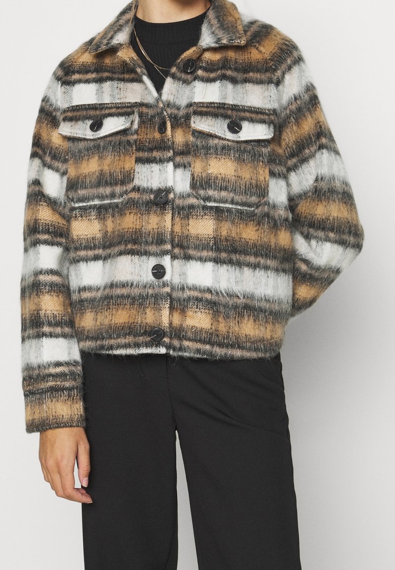Personne portant une veste à carreaux duveteuse en blanc, marron et gris avec des boutons noirs et un pantalon noir, debout devant un fond uni.