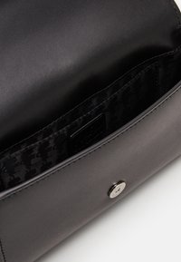 Musta nahkainen clutch-laukku, jossa on avonainen läppä, metallinen napitus ja kuvioitu kangasvuori, jossa on pieni tasku.