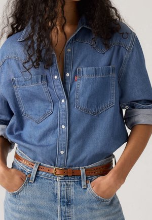 Kobieta nosząca rozpiętą niebieską koszulę dżinsową z podwiniętymi rękawami, brązowy skórzany pasek i jasnoniebieskie dżinsy, ręce w kieszeniach.