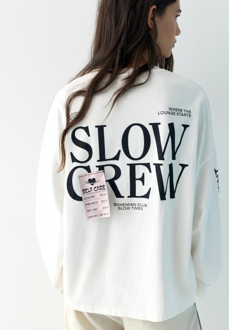 Ženska z dolgimi lasmi nosi belo preveliko srajco z napisom "SLOW CREW" in roza kontrolnim seznamom samopomoči na hrbtni strani.