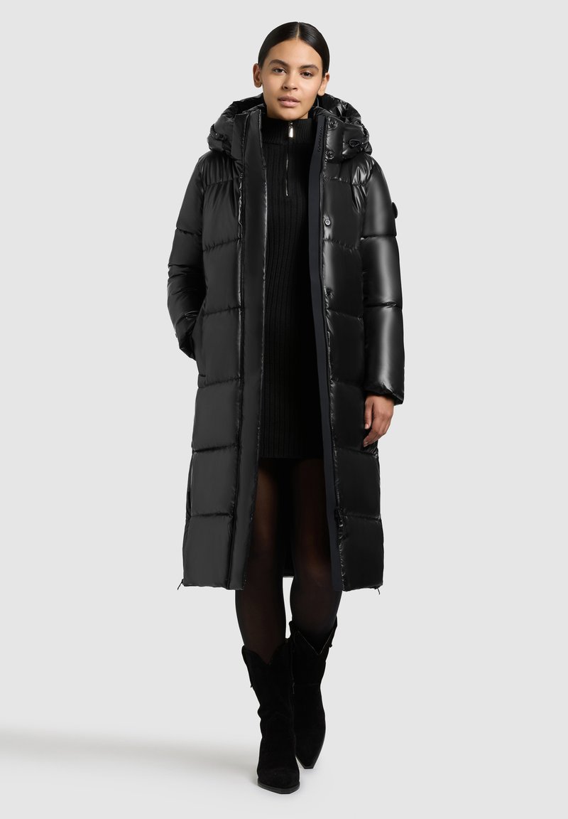 Abrigo acolchado largo, negro y con capucha, con secciones acolchadas y cierre de cremallera. Lleva puesto un vestido de suéter negro, combinado con botas negras.