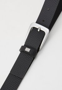 Ceinture en cuir noir avec une surface texturée, dotée d'une boucle en métal argenté et d'un passant en cuir avec logo. Design minimaliste sans motifs visibles.