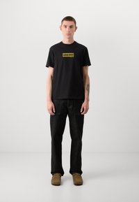 Svart t-skjorte med en gul Calvin Klein-logo, kombinert med svarte denim-bukser og brune semskede sko. Enkel, uformell design og passform.