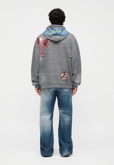 Άνδρας που φοράει γκρι φούτερ με ροζ φιόγκο και καρτούν σκύλους τυπωμένους στην πλάτη, σε συνδυασμό με φαρδιά ξεθωριασμένα μπλε τζιν, στέκεται με την πλάτη γυρισμένη.