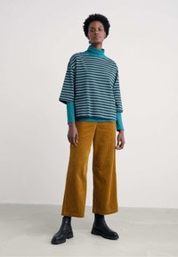 Nő, aki lazán áll, csíkos türkiz felsőt, mustár sárga széles szárú kordnadrágot és fekete bokacsizmát visel, egyszínű háttér előtt.