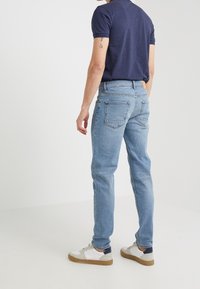 Muž v světle modrých džínách, námořnicko modré košili s krátkým rukávem a bílých teniskách stojí před čistě bílým pozadím.