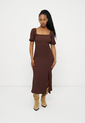 Hosszú fonott hajú nő barna midi ruhában, puffos ujjakkal és elöl hasítékkal, bézs félmagas csizmával, fehér háttér előtt állva.