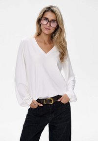 Weißes langärmliges V-Ausschnitt-Bluse mit gerafften Manschetten, kombiniert mit schwarzen hochgeschnittenen Hosen und einem braunen Gürtel mit einer goldenen Schnalle.