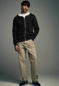 Must mustaga, millel on valge fliisvooder, on paaritatud beežide pükste ja mustade spordijalatsitega. Mudel seisab neutraalse tausta ees.