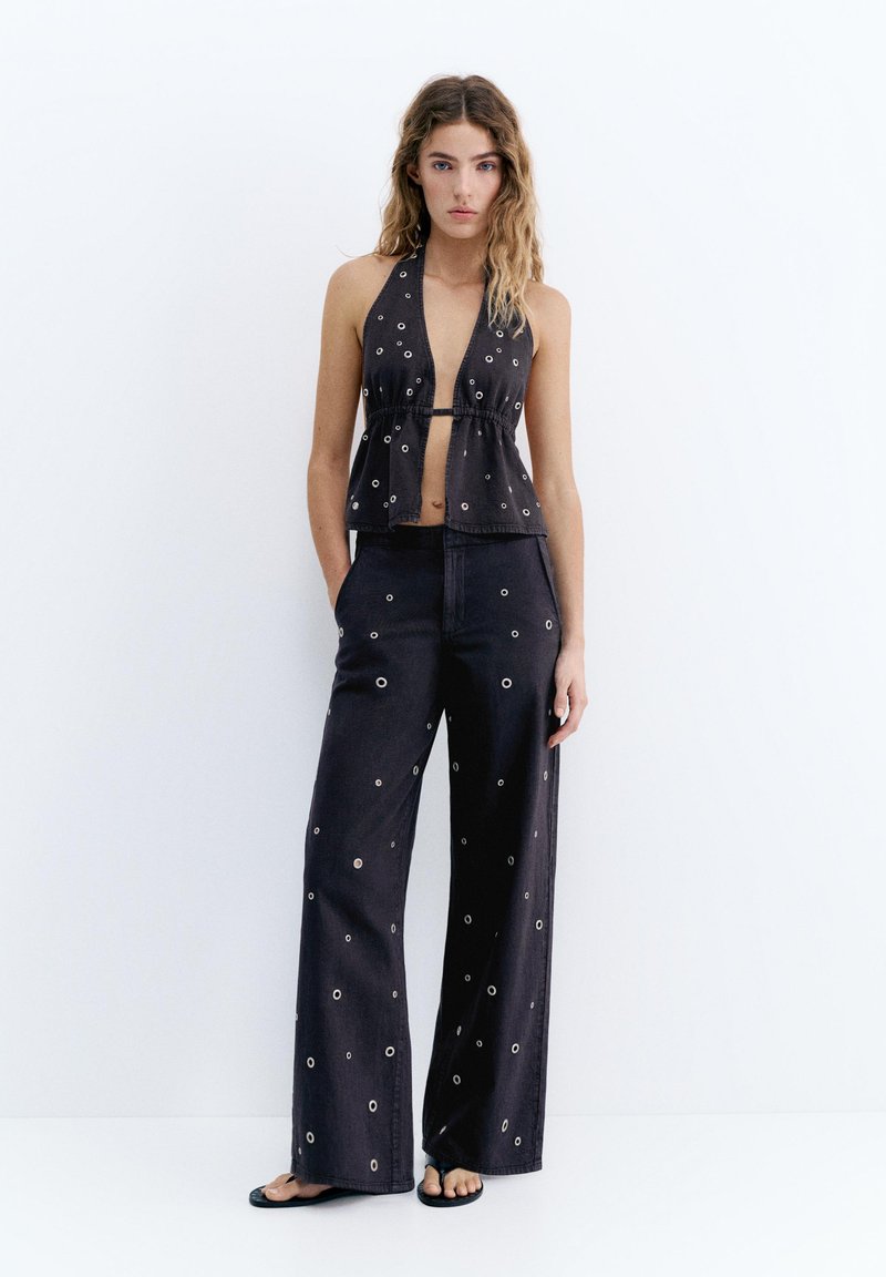 Jeune femme portant un top noir à col halter et un pantalon large avec des œillets argentés, se tenant devant un fond blanc uni.