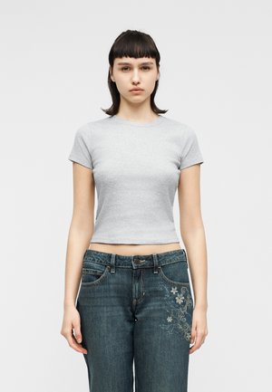 Noor naine sirgete mustade juustega kannab kitsast helehallikat kõhtu paljastavat toppi ja siniseid tikitud teksapükse, seistes ühevärvilise heleda tausta ees.