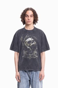 Fiatal személy göndör hajjal, elhasználódott fekete, oversize pólót visel, amelyen koponya és karom grafika található, valamint világoskék farmernadrágot.