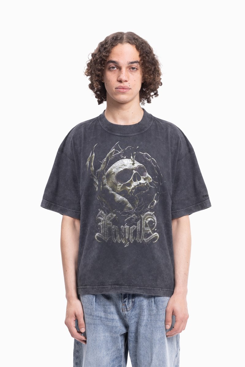 Fiatal személy göndör hajjal, elhasználódott fekete, oversize pólót visel, amelyen koponya és karom grafika található, valamint világoskék farmernadrágot.