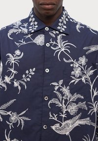 Laivastonsininen nappikauluspaita, jossa on monimutkainen valkoinen kukka- ja lintikirjonta, terävä kaulus ja erottuvat napit.