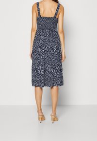 Navyblauwe jurk met bloemmotief, een smock-bustes, vierkante halslijn en gekruiste schouderbanden. Lengte tot halverwege de kuit en een gelaagde rok.