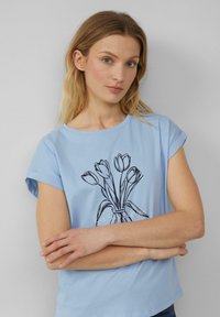 Hellblaues Baumwoll-T-Shirt mit aufgerollten Ärmeln, mit einer schwarzen Illustration eines Blumenvasens und dem Text "Rue des Fleurs" am unteren Rand.