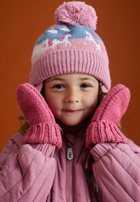 Różowe, dziergane rękawiczki oraz pasująca czapka z pomponem, ozdobione wzorami jednorożców i tęczy, noszone z różową kurtką puchową.