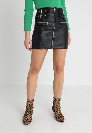 Vrouw draagt een zwarte leren minirok met voorrits en gespen, een groen geribd top met lange mouwen, en enkellaarsjes met luipaardprint.