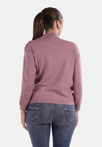 Donna con coda di cavallo castana che indossa orecchini a cerchio dorati, un maglione mauve e jeans blu, vista di spalle su uno sfondo neutro.