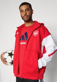 Red athletic jacket with white sleeves, featuring an Arsenal logo. The person holds a black and white soccer ball and wears a matching red shirt.