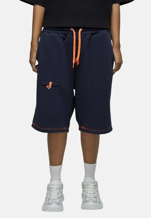 Personne portant un short bleu marine longueur genou avec des cordons oranges et un détail de poche, des chaussettes blanches et des chaussures de sport blanches.