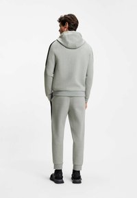 Lichtgrijze hoodie en joggingbroek van zachte stof met zwarte accenten aan de zijkanten. Heeft een capuchon en geribbelde manchetten, getoond vanaf de achterkant.