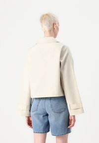Giacca beige tagliata con maniche ampie e colletto a punta. Indossata sopra pantaloni denim, mette in evidenza un materiale liscio e linee pulite.