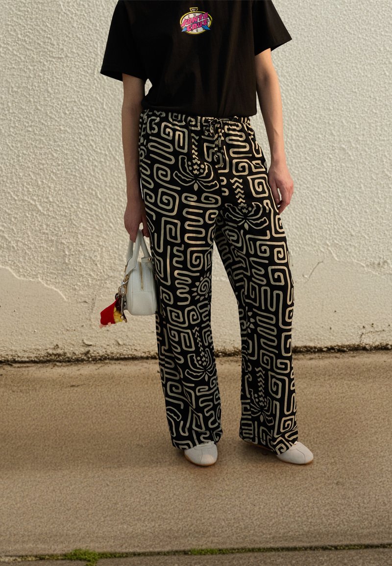 Παντελόνι με φαρδιά μπάρα σε μαύρο και λευκό μοτίβο με ρυθμιζόμενο κορδόνι στη μέση, συνδυασμένο με μαύρο μπλουζάκι και λευκή τσάντα χεριού. Ορατά παπούτσια με επίπεδη σόλα.