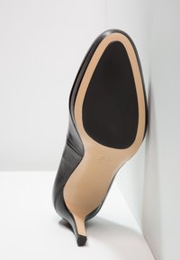 Chaussure à talon haut en cuir noir avec semelle beige et semelle en caoutchouc noire, positionnée à la verticale contre un mur blanc et une surface blanche.