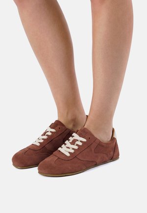 Sneakers in camoscio marrone con lacci bianchi indossate a piedi nudi, viste da angoli laterali e frontali su uno sfondo chiaro uniforme.