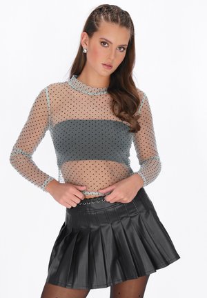 Ung kvinde med fletninger iført en gennemsigtig, langærmet top med prikker over en grå tube top og en sort plisseret mininederdel.