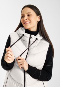 Witte gewatteerde vest met capuchon, zwarte accenten en ritsluiting. Beschikt over een gequilt ontwerp en een trekkoord bij de halslijn. Zwarte trui eronder.