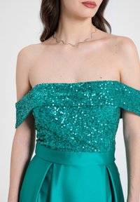 Abito turchese senza spalline con un corpetto di paillettes e gonna di raso. Caratterizzato da una collana d'argento ondulata. Texture liscia, finitura lucida.