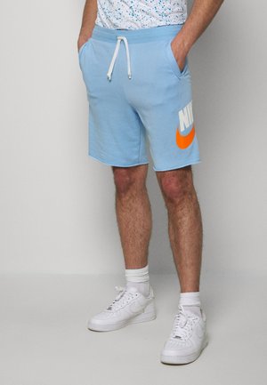 Man draagt lichtblauwe Nike-shorts met een witte trekkoord, witte sokken en witte sneakers, staat met handen in de zakken.