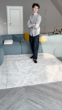 Sofá de tecido azul claro com design curvo, combinado com um tapete felpudo branco. O chão apresenta um padrão de espinha de peixe em tons de madeira cinza.
