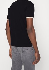 Man i svart kortärmad skjorta med randiga manschetter och blekta grå jeans, sedd bakifrån mot en enfärgad bakgrund.