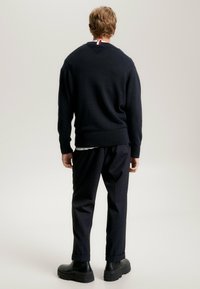 Pull bleu marine foncé, poignets et ourlet côtelés, col en V avec des accents rouges et blancs à l'arrière ; associé à un pantalon noir texturé et des chaussures noires épaisses.