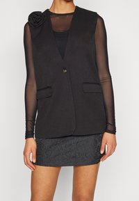 Černá vesta s jedním knoflíkem, dvěma předními kapsami a růžovým akcentem na levém rameni, doplněná o texturovanou černou sukni.