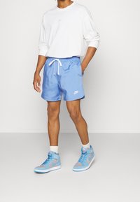 Osoba v bílém tričku s dlouhým rukávem Nike, světle modrých šortkách Nike s tkaničkou, bílých ponožkách a modrošedých vysokých teniskách Nike stojí proti jednoduchému pozadí.