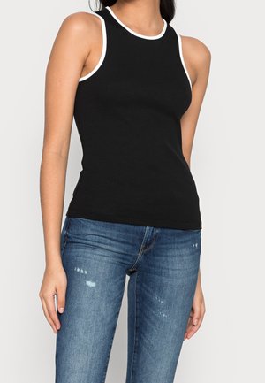 Vrouw draagt een mouwloze zwarte top met witte bies, gecombineerd met blauwe versleten skinny jeans, staand tegen een effen achtergrond.