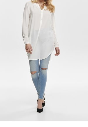 Blouse blanche à manches longues avec col et boutons, associée à un jean skinny bleu clair déchiré aux genoux, et des escarpins noirs à bouts pointus.